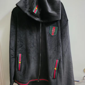 Svart tracksuit med gröna och röda detaljer - Säljer en svart tracksuit med mjuk finish, gröna och röda ränder samt broderade detaljer med gul stjärna. Jackan har huva, dragkedja och emblem på bröstet. Byxorna har snörning i midjan och matchande ränder vid fickor och benslut. Perfekt för chill eller streetwear.