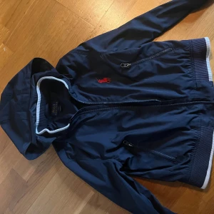 Marinblå vindjacka Polo Ralph Lauren  - Marinblå vindjacka från Polo Ralph Lauren med huva, dragkedja och ribbade muddar. Jackan har röd broderad logga på bröstet och två fickor med dragkedja. Tillverkad i polyester, perfekt för blåsiga dagar. För barn.