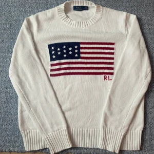 Vit stickad tröja Polo Ralph Lauren - Vit stickad tröja från Polo Ralph Lauren med amerikansk flagga i blått, rött och vitt på bröstet samt RL-broderi. Rund hals och ribbade muddar vid ärmslut och nederkant. Perfekt för dig som gillar klassisk och preppy stil. Ställ gärna frågor eller lägg bud😁