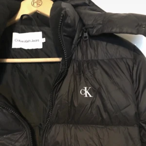 Svart pufferjacka Calvin Klein Jeans M - Svart pufferjacka från Calvin Klein Jeans med broderad cK-logga på bröstet. Jackan har huva, dragkedja och är tillverkad av återvunnen nylon och polyester med dunfyllning. Vattenavvisande och perfekt för kyliga dagar.