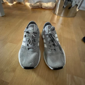 Grå Adidas sneakers med vita ränder - Säljer ett par grå Adidas sneakers med klassiska vita ränder på sidorna och vit sula. Ovandelen är i mesh-liknande syntetmaterial som andas och har snörning framtill. Perfekta för dig som gillar en clean och sportig look. En liten defekt på en sko som var ett hål som sedan blev sytt ihop med kvalité! 