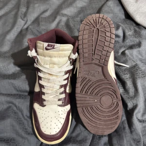 Nike Dunk High vinröd och vit - Säljer ett par Nike Dunk High sneakers i vinrött och vitt. Skorna har klassisk hög siluett, snörning och rund tå. Ovandelen är i skinn med perforerade detaljer på tån och Nike-logga på plösen. Perfekta för dig som gillar retrostil och streetwear. Storlek 42. Dom ska bort