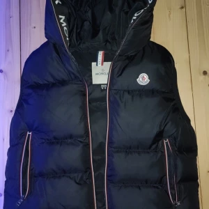 Svart dunväst från Moncler - Svart dunväst från Moncler med huva och klassisk logga på bröstet. Västen har två fickor med dragkedja och snygga röda och vita detaljer längs dragkedjorna. Perfekt för lager-på-lager och streetstyle.