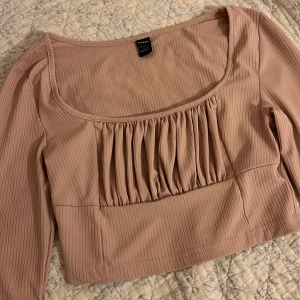 Ribbad rosa croppad topp från SHEIN - Säljer en ljusrosa croppad topp från SHEIN med långa ärmar och ribbad struktur. Toppen har en snygg rynkad detalj framtill och bred, fyrkantig urringning. 