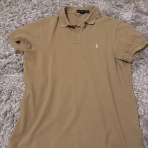 Beige pikétröja från Polo Ralph Lauren - Klassisk beige pikétröja från Polo Ralph Lauren med broderad logga på bröstet. Tröjan har korta ärmar, krage och knäppning framtill. Perfekt för en clean och stilren look.