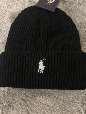 Svart stickad mössa från Polo Ralph Lauren - Svart ribbstickad mössa från Polo Ralph Lauren med klassisk broderad logga i vitt framtill. Mössan är tillverkad i mjukt material och har en bred uppvikt kant för extra stil. Perfekt för att hålla värmen och se snygg ut under kyliga dagar.