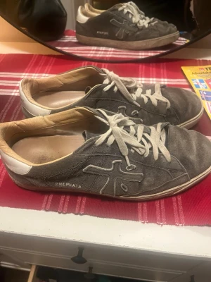 Premiata grå sneakers med mocka - Säljer ett par grå sneakers från Premiata med vit häl och beige insida. Skorna har snörning och är tillverkade i mocka med detaljerad logga på sidan. Sulan är platt och har Premiata-märkning undertill. Perfekt för dig som gillar stilrena och klassiska sneakers.