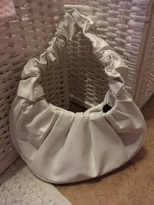 Vit axelväska med rynkad rem - Säljer en superfin vit axelväska med rynkad rem och mjuk, paljettliknande form. Väskan har magnetknäppning och är tillverkad i syntetmaterial. Perfekt för dig som vill ha en trendig accessoar med unik design.