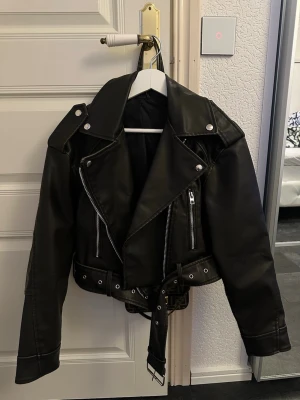 Armegrön skinnjacka med bälte och dragkedjor - Snygg armegrön skinnjacka med coola silverfärgade dragkedjor och detaljer. Jackan har ett brett bälte med metallspänne, klassisk krage och långa ärmar. Croppad passform som ger en edgy vibe. Perfekt för dig som vill ha en stilren och tuff look.