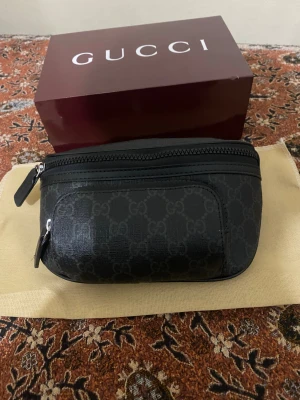Svart Gucci axelväska med GG-mönster - Snygg svart axelväska från Gucci med klassiskt GG-mönster över hela väskan. Väskan har justerbart band, dragkedja upptill och en extra ficka framtill. Materialet är skinn och detaljerna är i svart. Perfekt för dig som vill ha en stilren och lyxig look.