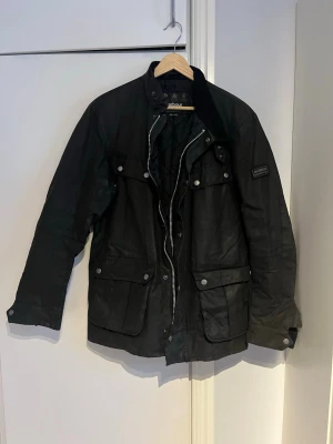 Svart Barbour International jacka M - Svart Barbour International jacka med quiltad insida, flera fickor med tryckknappar och dragkedja framtill. Yttertyget är vaxad bomull, fodret är polyamid och fyllningen polyester. Klassisk look med ståkrage och Barbour-logga på ärmen. Står emot regn.