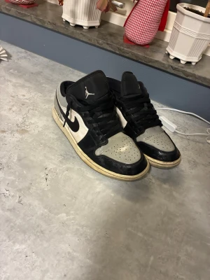 Nike Jordan 1 Low svart/grå sneakers - Nike Jordan 1 Low sneakers i svart, grått och vitt med klassisk swoosh på sidan och Air Jordan-logga på hälen. Skorna har snörning, perforerad tå och platt sula. Materialet är skinn och syntet, och modellen är lows vilket ger en schysst streetstil.