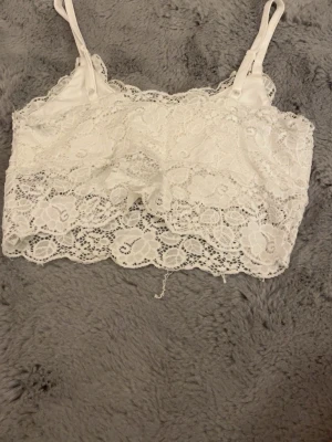 Vit spetsig croppad linnetopp - Säljer en vit croppad linnetopp med justerbara axelband och vacker spets över hela toppen. Blommigt spetsmönster ger en romantisk vibe och toppen är ärmlös med en feminin siluett. Perfekt att styla med jeans eller kjol för en fräsch look. Kan eventuellt tänka mig gå ner i pris