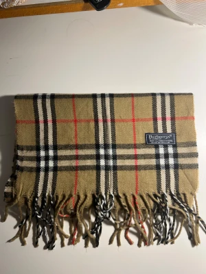 Klassiskt halsduk från Burberry - Säljer en klassisk halsduk från Burberrys i beige med det ikoniska rutiga mönstret i svart, vitt och rött. Västen är tillverkad i 100% lammull och har fransar längst ner. Perfekt för dig som gillar tidlös design och vill sticka ut med ett lyxigt plagg.