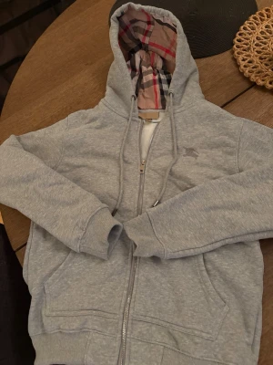 Grå Burberry Hoodie  - Säljer en grå hoodie från Burberry i storlek M. Tröjan har dragkedja, snörning vid huvan och Burberrys klassiska rutiga mönster på insidan av huvan. Broderad logga på bröstet och två fickor framtill. Perfekt för dig som vill ha en stilren och lyxig look. Passar även S