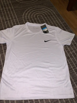 Vit Nike Dri-Fit t-shirt herr M - Snygg vit Nike Dri-Fit t-shirt med svart swoosh-logga på bröstet. Tillverkad i lätt och ventilerande polyester som passar perfekt för fotboll eller träning. Rund hals och korta ärmar, stilren och sportig design. Taggen sitter kvar.
