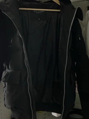 Svart pufferjacka Supply & Demand S - Svart pufferjacka från Supply & Demand med huva, stora fickor och coola dragkedjor. Jackan har en snygg patch på ärmen och är perfekt för kalla dagar. Stilen är modern och passar dig som gillar streetwear.