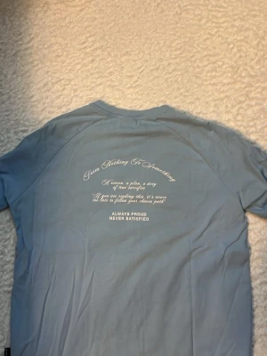 Blå HOODRICH t-shirt med tryck - Snygg ljusblå t-shirt från HOODRICH med stort vitt tryck på bröstet och textdetaljer både fram och bak. T-shirten har en klassisk rund hals och korta ärmar. Materialet är mjuk bomull som känns skönt mot huden. Perfekt för dig som gillar streetwear och vill sticka ut. Ny t shirt.