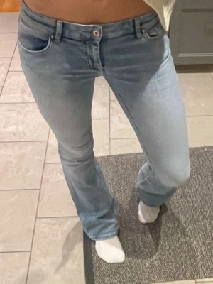 Bootcut ljusblå jeans - Säljer ett par ljusblå bootcut jeans med klassisk femficksdesign och knappgylf. Jeansen har en snygg, något utsvängd passform nedtill och är tillverkade i mjukt denimtyg som sitter skönt. Perfekta för dig som gillar retrovibbar och vill ha en cool look.