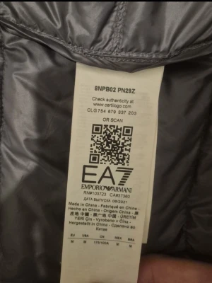 Grå pufferjacka från Emporio Armani EA7 - Säljer en grå pufferjacka från Emporio Armani EA7 med huva och dragkedja framtill. Jackan har quiltad design och logga på bröstet. Perfekt för dig som vill ha en snygg och lätt jacka med stilren look. Materialet är polyester och jackan är långärmad.