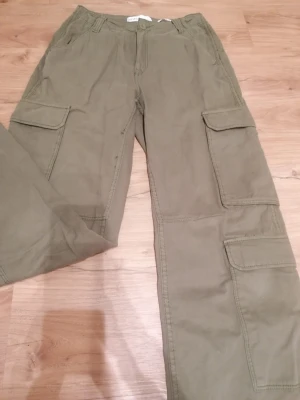 Khakigröna cargopants från Bershka - Säljer ett par khakigröna cargopants från Bershka med flera praktiska fickor på benen och klassisk dragkedja framtill. Byxorna har en rak passform och är tillverkade i bomullsmaterial. Perfekt för dig som gillar streetstyle och funktionella detaljer. De är endast använda ett fåtal gånger! 😊
