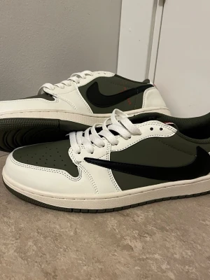 Nike Air Jordan 1 Low Travis Scott - Nike Air Jordan 1 Low Travis Scott i olivgrönt och vitt skinn med svart swoosh och röda broderade detaljer på hälen. Klassisk rund tå, vita snören och grön yttersula. Snygg streetwear-vibe och ikonisk design som sticker ut.