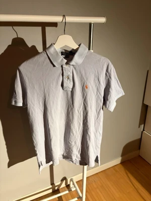 Vit kortärmad piké från Polo Ralph Lauren - Säljer en klassisk vit pikétröja från Polo Ralph Lauren med korta ärmar och krage. Tröjan har två knappar framtill och den ikoniska broderade loggan i orange på bröstet. Tillverkad i mjuk bomull som är skön mot huden.