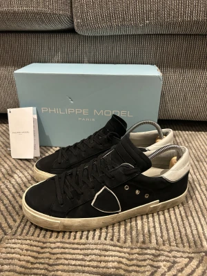Svarta Philippe Model sneakers - Snygga svarta sneakers från Philippe Model med vita detaljer på hälen och en vit kant runt sulan. Skorna har snörning, rund tå och platt sula. Materialet är skinn och textil, med metallnitar på sidan och en ikonisk sköldpatch. Perfekt för dig som gillar stilrena och trendiga sneakers.