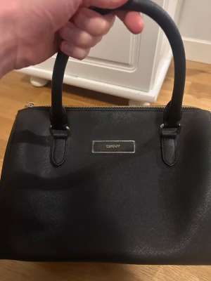 Svart handväska från DKNY i skinn - Dkny väska i mycket fint skick. Äkta läder. Det enda anmärkningsbara är att den har blivit lite ihoptryckt men det borde man kunna åtgärda enkelt.  Mått: 30 cm lång 12 cm bred och 23 cm hög.