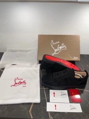 Svarta sneakers från Christian Louboutin - Snygga svarta sneakers från Christian Louboutin med klassisk röd sula och broderad logga på hälen. Skorna har rund tå, platt sula och är tillverkade i mjuk mocka med svarta snören. Perfekt för dig som gillar exklusiv och stilren design.
