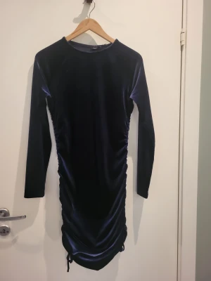 Marinblå sammetsklänning med snörning - Säljer en marinblå sammetsklänning med lång ärm och snygg snörning längs sidorna. Klänningen har en tight passform och är kort i modellen, vilket ger en cool och trendig look. Perfekt för dig som gillar mjuka material och stilrena färger.