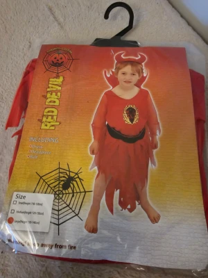 Röd djävulsväst barn Large - Säljer en röd väst för barn med eldigt tryck framtill och svarta detaljer. Västen ingår i ett djävulskostymset och har en cool, taggig nederkant. Perfekt för halloween eller maskerad! Huvudbonad och bälte medföljer.