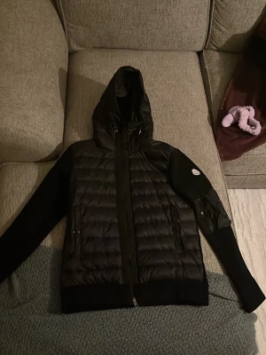 Svart Moncler pufferjacka med huva - Svart pufferjacka från Moncler med quiltad framsida och huva. Jackan har dragkedja, ribbade muddar och fickor med dragkedja. Ärmarna är släta och har en Moncler-logga på vänster arm. Perfekt för dig som vill ha en snygg och varm jacka med exklusiv känsla.
