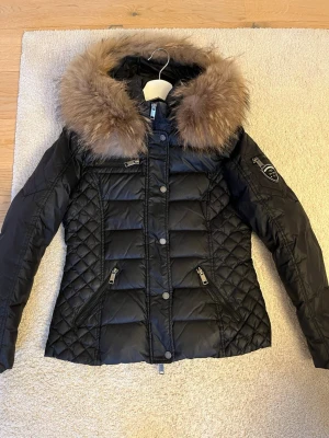 Svart dunjacka Rockandblue med päls - Snygg svart dunjacka från Rockandblue med fluffig päls runt kragen. Jackan har quiltade detaljer på sidorna och ärmarna, dragkedjor på fickorna och en cool patch på ena ärmen. Perfekt för kalla dagar och har en riktigt schysst look. En liten skada på ena armen, se bild. Går nog att sy ihop enkelt om man skulle vilja. Liten i storlek, passar även stl 36.