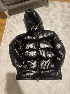 Svart pufferjacka/vinterjacka från Moncler - Säljer en svart, glansig pufferjacka/vinterjacka från Moncler med huva och dragkedja. Jackan har en klassisk Moncler-logga på ärmen och är fylld med dun för extra värme. Perfekt för kalla dagar och riktigt snygg streetstil. Priset kan diskuteras
