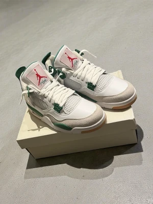 Nike SB Air Jordan 4 vit/grön sneakers - Nike SB Air Jordan 4 sneakers i vitt och grönt med röda detaljer på tungan och yttersulan. Skorna har meshpaneler, snörning och en platt gummisula med brun detalj. Ovandelen är i skinn och mocka, med klassisk Air Jordan-logga och Nike SB branding på hälen. (Obs ej original)
