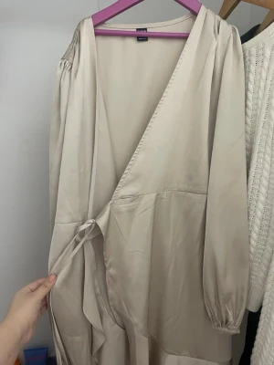 Beige-grön omlottklänning från SHEIN  - Säljer en beige-grön omlottklänning från SHEIN med långa ärmar och knyt i midjan. Klänningen har en v-ringad design och är tillverkad i ett mjukt, slätt material som ger en elegant känsla. Perfekt för dig som gillar stilrena och enkla plagg.