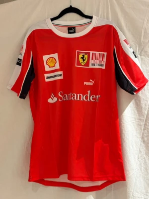 Ferrari f1 2010 tröja - Röd Ferrari x Puma-tröja från 2010-eran, året då Ferrari slogs om VM-titeln med Fernando Alonso. Klassiska sponsors som Santander, Shell och Bridgestone – exakt den looken man förknippar med Ferrari runt 2010.  Tröjan är i fint skick och funkar lika bra som vardagsplagg som för F1- eller Ferrari-fans. Skriv om du vill ha fler bilder eller mått. 