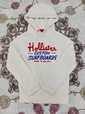 Vintage Hollister Hoodie Custom Surfboards Malibu - Snygg vit hoodie från Hollister med trycket Custom Surfboards Made in Malibu i rött och blått. Klassisk surf/skater-stil som passar till allt. Mjuk och bekväm med huva och ficka framtill.  Längd från axeln och ner: 61 cm  Bredd från axel till axel: 40 cm Längden på armarna:  59 cm  📦 Skickas snabbt & välpackat.