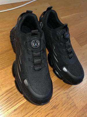 Svarta sneakers med snörning - Säljer ett par svarta sneakers från SafeLand i storlek 42.5. Skorna har en chunky sula med rundad form och detaljer i grått. Ovandelen är gjord av mesh och syntetmaterial, med snörning och en rund tå. Perfekta för dig som gillar sportig stil.
