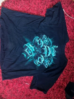 Svart t-shirt med rockigt döskalletryck - Cool svart t-shirt med stort grafiskt tryck av döskallar och texten 'Rock Skulls' i turkos. T-shirten har korta ärmar och en klassisk rund hals. Perfekt för dig som gillar rock och vill sticka ut med en edgy stil. Materialet känns som mjuk bomull.