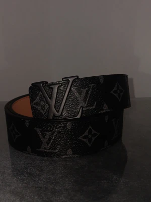 Louis Vuitton svart monogram skärp - Säljer ett svart Louis Vuitton skärp med klassiskt monogrammönster och stor LV-logga i metall på spännet. Utsidan är svart med grått mönster, insidan är ljusbrun och skärpet är gjort i skinn. Perfekt accessoar för att lyfta din outfit.