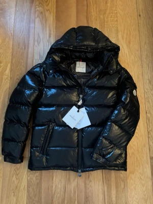 Svart pufferjacka från Moncler - Svart pufferjacka från Moncler med glansig finish och huva. Jackan har dragkedja framtill, två sidofickor med dragkedja och ett coolt serietryck på insidan. Perfekt för dig som vill ha en varm och snygg jacka med streetkänsla.