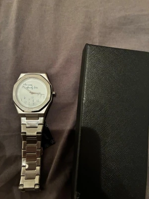 Silverfärgad klocka från Miler - Säljer en stilren klocka från Miler i silverfärgat rostfritt stål. Urtavlan har ett unikt och lekfullt tryck med texten 'Who cares I'm already late' och siffrorna är placerade huller om buller. Länken är klassisk och boetten har en kantig form som ger en modern vibe.