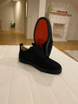 Svarta sneakers från Christian Louboutin. - Snygga svarta sneakers från Christian Louboutin med klassisk röd sula och diskret logga. Skorna har rund tå, snörning och är tillverkade i mocka med beige innersida. Perfekt för dig som gillar stilrena och lyxiga detaljer. Ingen kartong eller kvitto finns med.