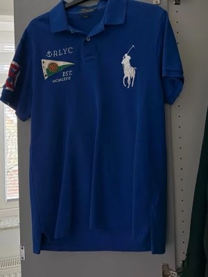 Blå pikétröja Polo Ralph Lauren - Säljer en blå pikétröja från Polo Ralph Lauren i slim fit. Tröjan har korta ärmar, klassisk krage och knappar framtill. På bröstet finns ett stort broderat polospelare-motiv och flera broderade detaljer, bland annat flagga och text. Perfekt för dig som gillar sportig stil.