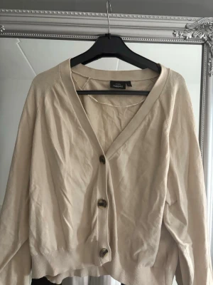 Beige kofta från KappAhl med knappar - Beige kofta från KappAhl med v-ringad hals och tre stora knappar framtill. Modellen är croppad med ribbade muddar vid nederkant och ärmslut. Storleken är L men den är ganska liten i storlek