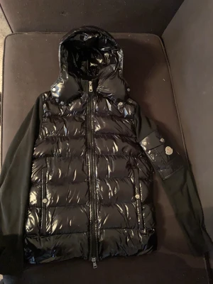 Moncler Cardigan Vintage - Säljer en svart dunväst från Moncler med glansig yta och avtagbar huva. Västen har dragkedja framtill, två fickor med tryckknappar och ett Moncler-märke på ärmen. Materialet är polyester och dun, med stickade partier på sidorna och ryggen för extra komfort.