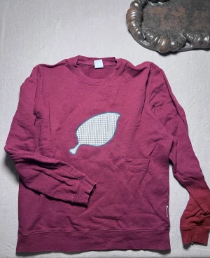 Röd sweatshirt från nno7 - Säljer en röd sweatshirt med ett stort tryck i rutigt mönster på bröstet. Tröjan har rund hals, långa ärmar och är gjord i mjuk bomull. Perfekt för dig som gillar unika prints och vill ha en chill stil.