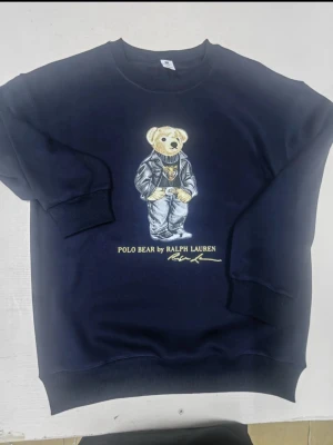 Mörkblå sweatshirt Polo Bear Ralph Lauren - Säljer en mörkblå sweatshirt från Ralph Lauren med Polo Bear-tryck på bröstet. Tröjan har rund hals, ribbade muddar och en loose passform. Materialet är mjuk bomull och motivet föreställer en nalle i cool outfit. Perfekt för dig som gillar streetwear och snygga prints.